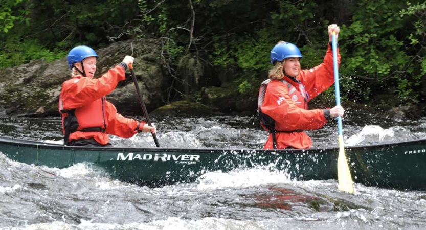 Two people wearing safety gear paddle a canoe through choppy water